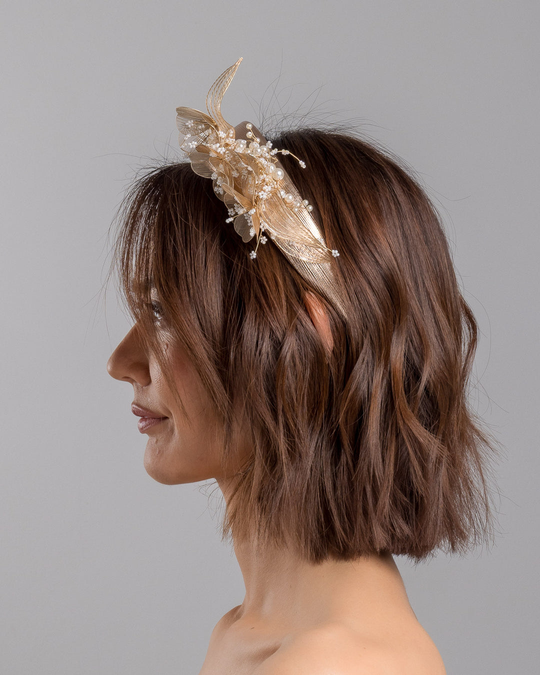 The model is showcasing the left side close up of the gold metallic headband with sculpted floral and leaf embellishments, featuring delicate ivory bead details and an elegant asymmetrical design. Designed and made by Merve Bayindir Millinery.
