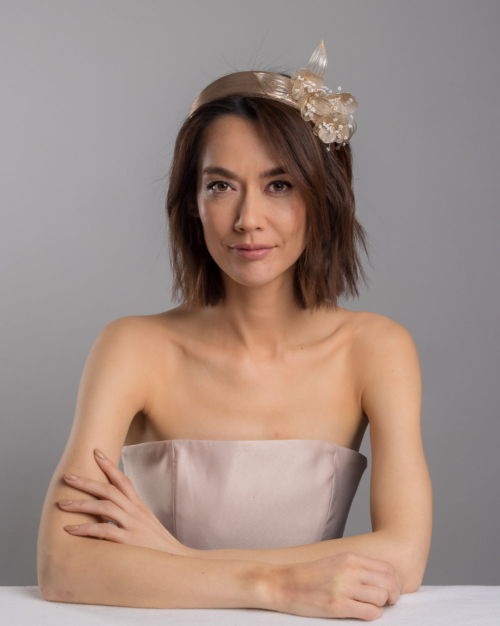 The female model is posing, wearing Alis,  the gold metallic headband with sculpted floral and leaf embellishments, featuring delicate ivory bead details and an elegant asymmetrical design. Designed and made by Merve Bayindir Millinery.
