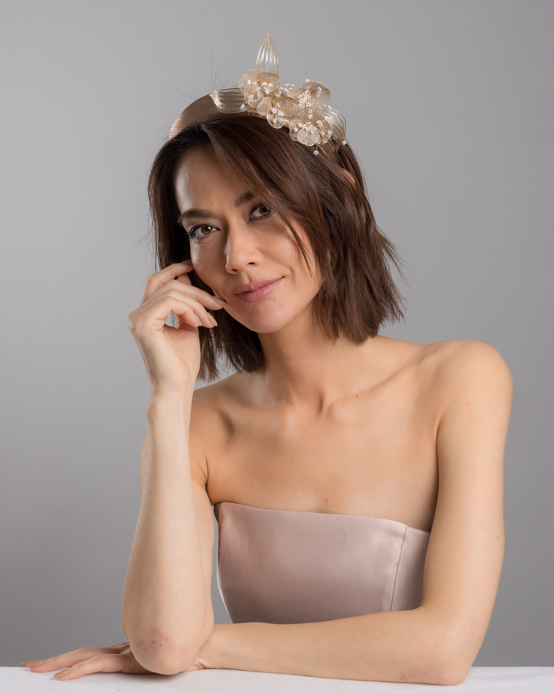 The female model is posing, wearing Alis,  the gold metallic headband with sculpted floral and leaf embellishments, featuring delicate ivory bead details and an elegant asymmetrical design. Designed and made by Merve Bayindir Millinery.
