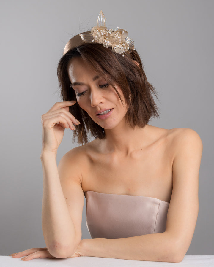 The female model is posing, wearing Alis,  the gold metallic headband with sculpted floral and leaf embellishments, featuring delicate ivory bead details and an elegant asymmetrical design. Designed and made by Merve Bayindir Millinery.
