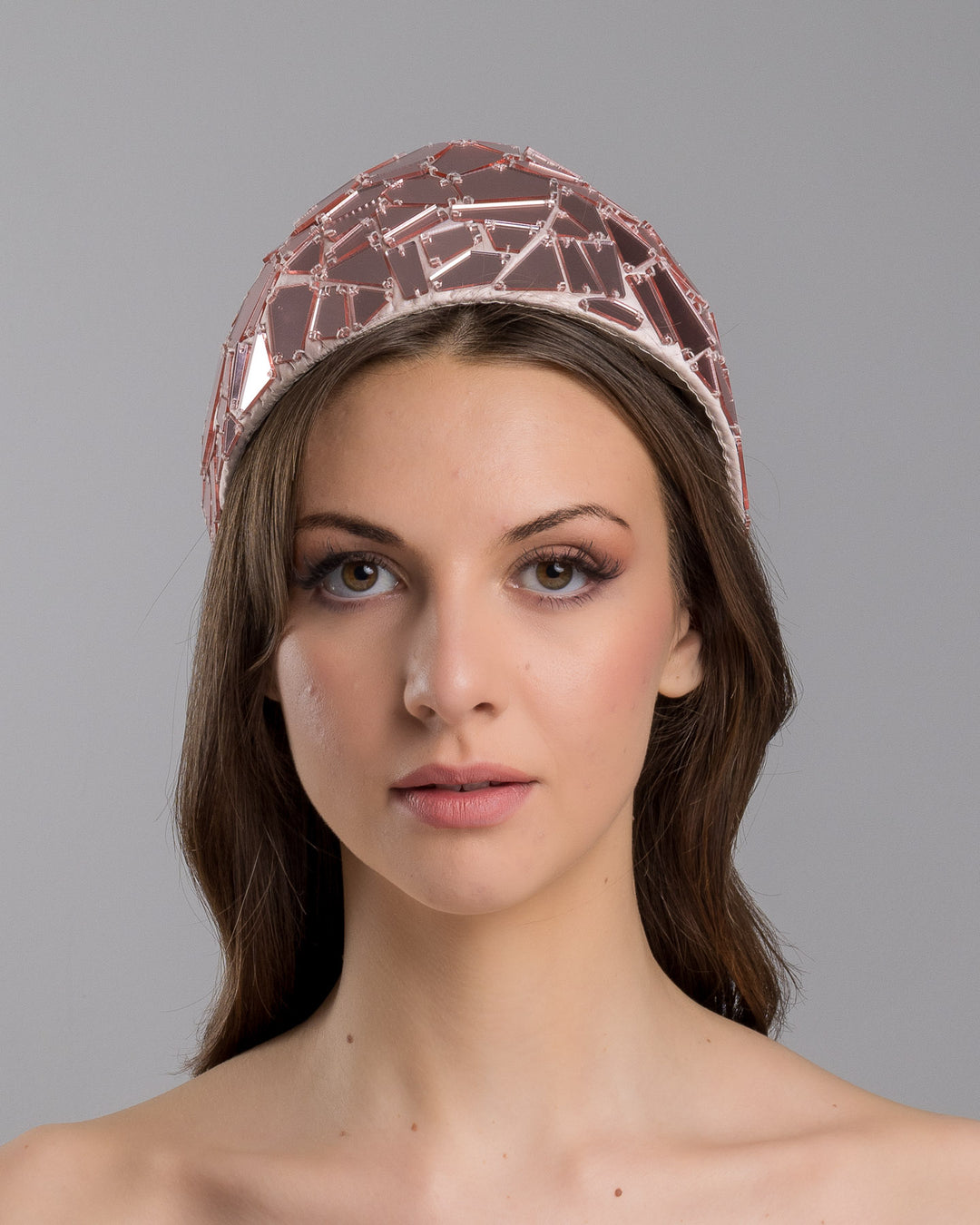 The model is showcasing the front side close up of the blush-toned structured half-hat embellished with geometric mirrored panels in soft rose and clear finishes, arranged in a mosaic pattern and lined with smooth champagne satin. Designed and made by Merve Bayindir Millinery.

