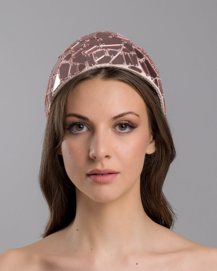 The model is showcasing the front side close up of the blush-toned structured half-hat embellished with geometric mirrored panels in soft rose and clear finishes, arranged in a mosaic pattern and lined with smooth champagne satin. Designed and made by Merve Bayindir Millinery.
