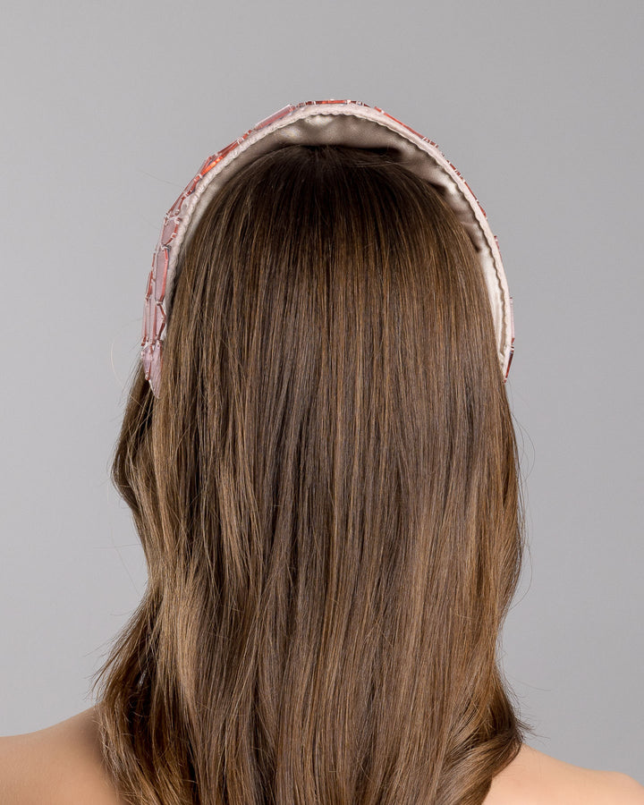 The model is showcasing the back side close up of the blush-toned structured half-hat embellished with geometric mirrored panels in soft rose and clear finishes, arranged in a mosaic pattern and lined with smooth champagne satin. Designed and made by Merve Bayindir Millinery.
