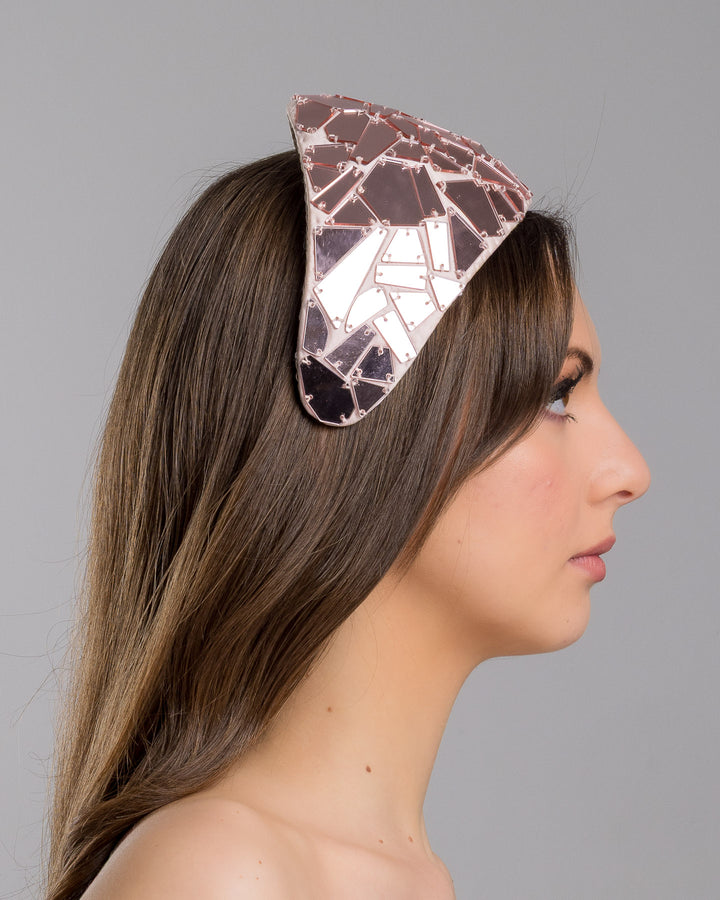 The model is showcasing the right side close up of the blush-toned structured half-hat embellished with geometric mirrored panels in soft rose and clear finishes, arranged in a mosaic pattern and lined with smooth champagne satin. Designed and made by Merve Bayindir Millinery.

