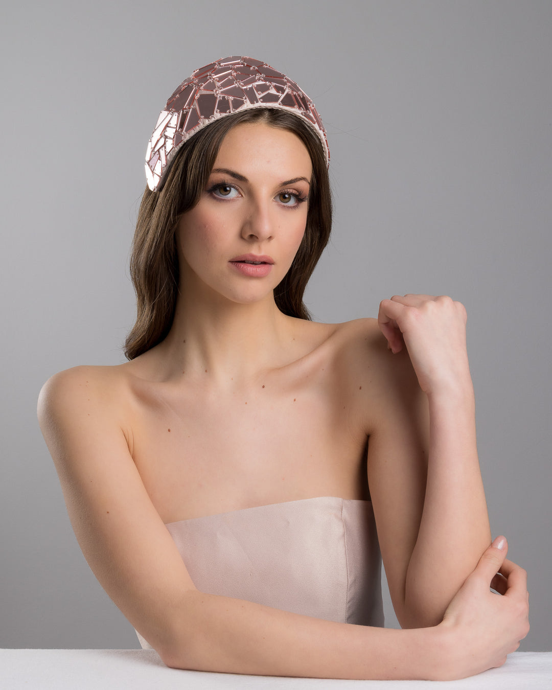 The female model is posing, wearing
Anna, the blush-toned structured half-hat embellished with geometric mirrored panels in soft rose and clear finishes, arranged in a mosaic pattern and lined with smooth champagne satin. Designed and made by Merve Bayindir Millinery.
