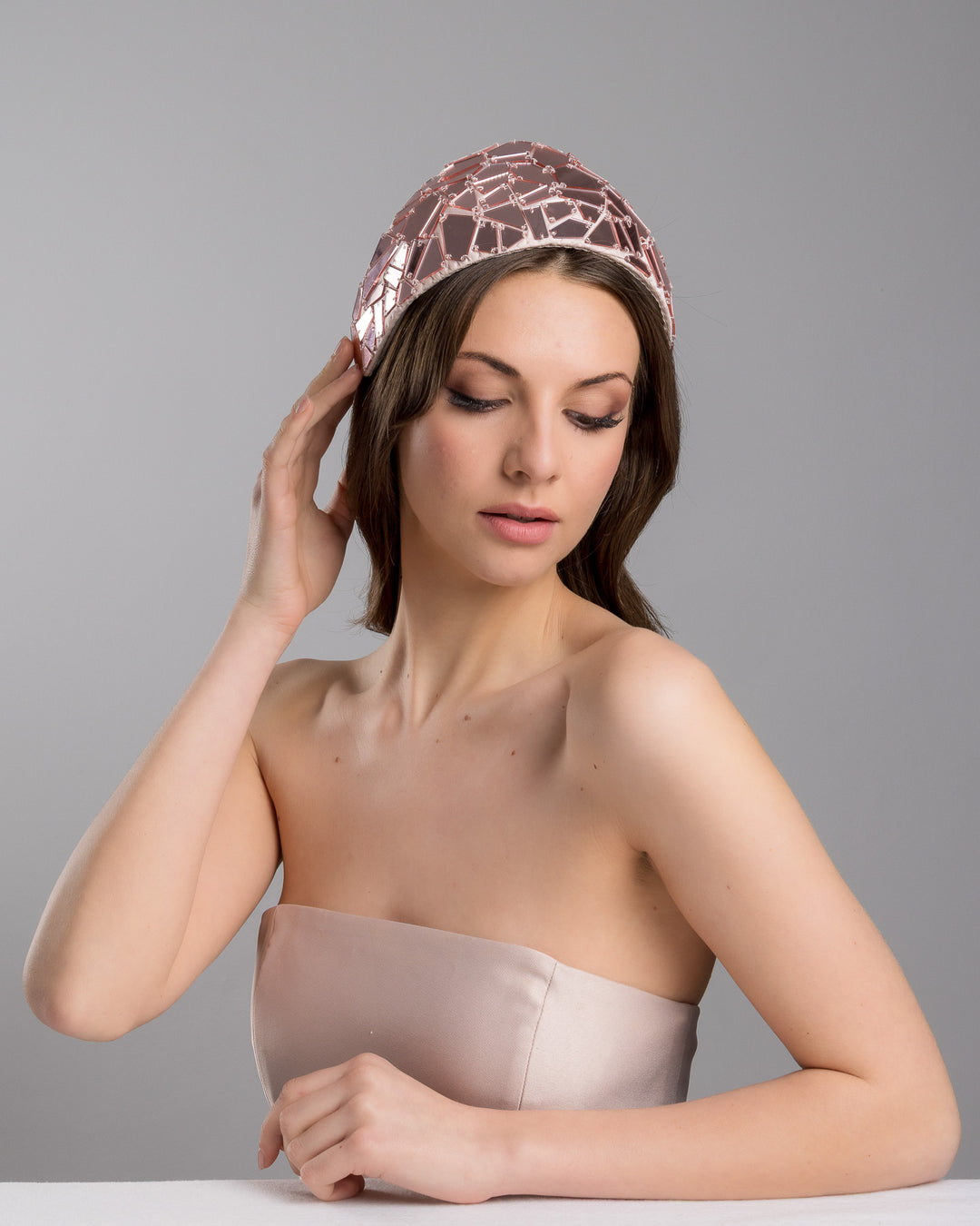 The female model is posing, wearing
Anna, the blush-toned structured half-hat embellished with geometric mirrored panels in soft rose and clear finishes, arranged in a mosaic pattern and lined with smooth champagne satin. Designed and made by Merve Bayindir Millinery.

