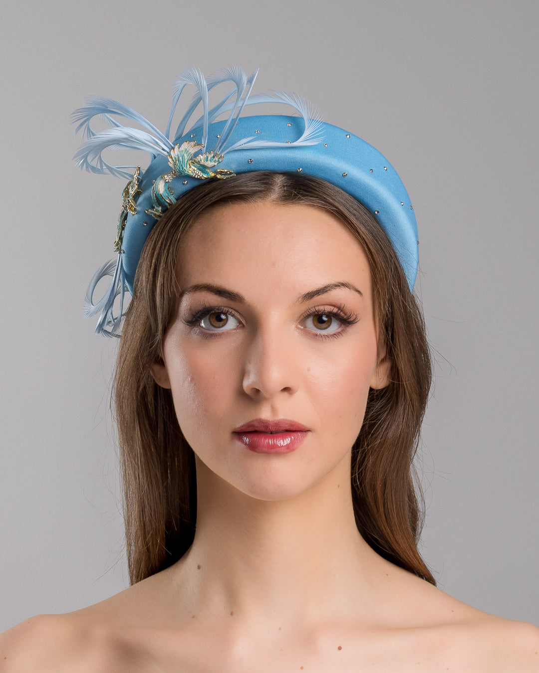 The model is showcasing the front side close up of the aqua blue padded headband embellished with crystal accents, featuring gold and blue bird appliqués with flowing feather details for a bold, statement finish. Designed and made by Merve Bayindir Millinery.
