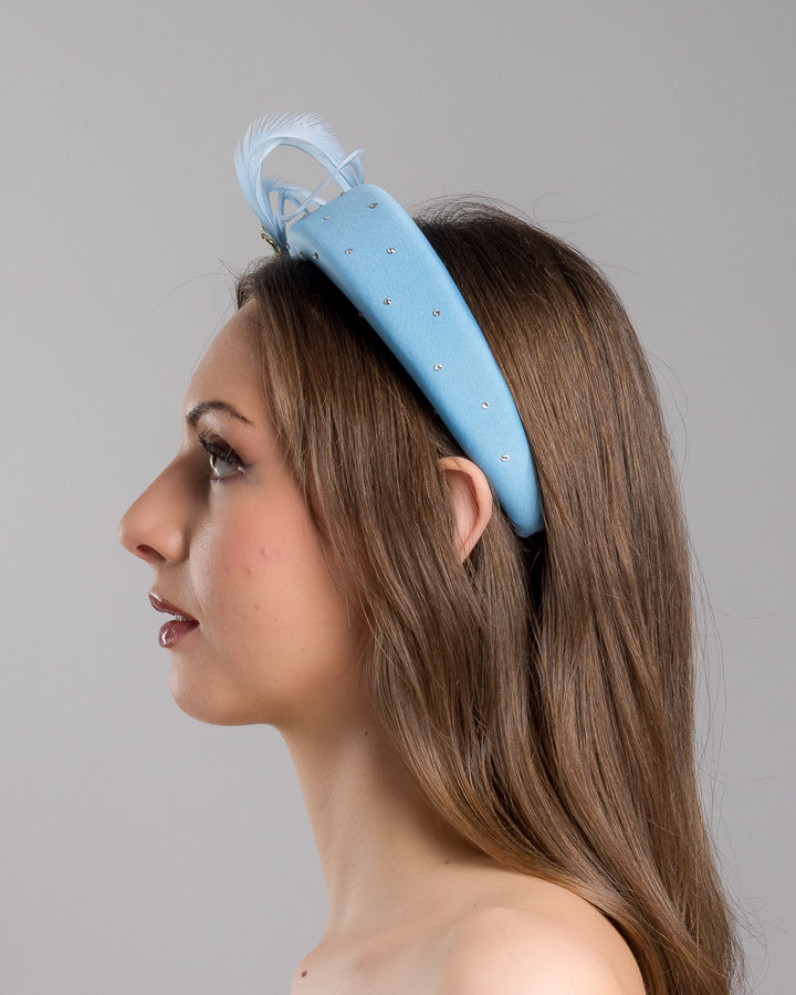 The model is showcasing the left side close up of the aqua blue padded headband embellished with crystal accents, featuring gold and blue bird appliqués with flowing feather details for a bold, statement finish. Designed and made by Merve Bayindir Millinery.
