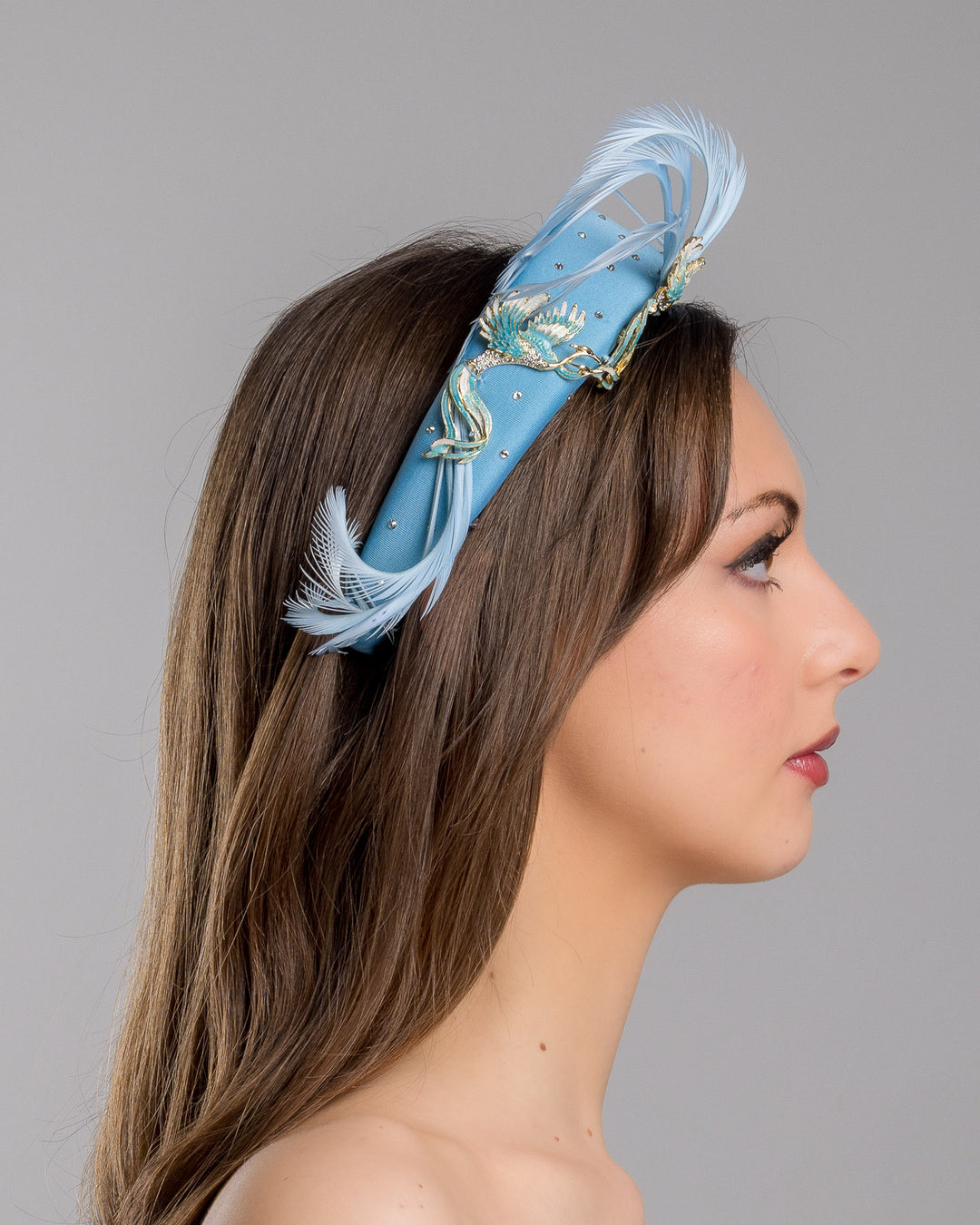 The model is showcasing the right side close up of the aqua blue padded headband embellished with crystal accents, featuring gold and blue bird appliqués with flowing feather details for a bold, statement finish. Designed and made by Merve Bayindir Millinery.

