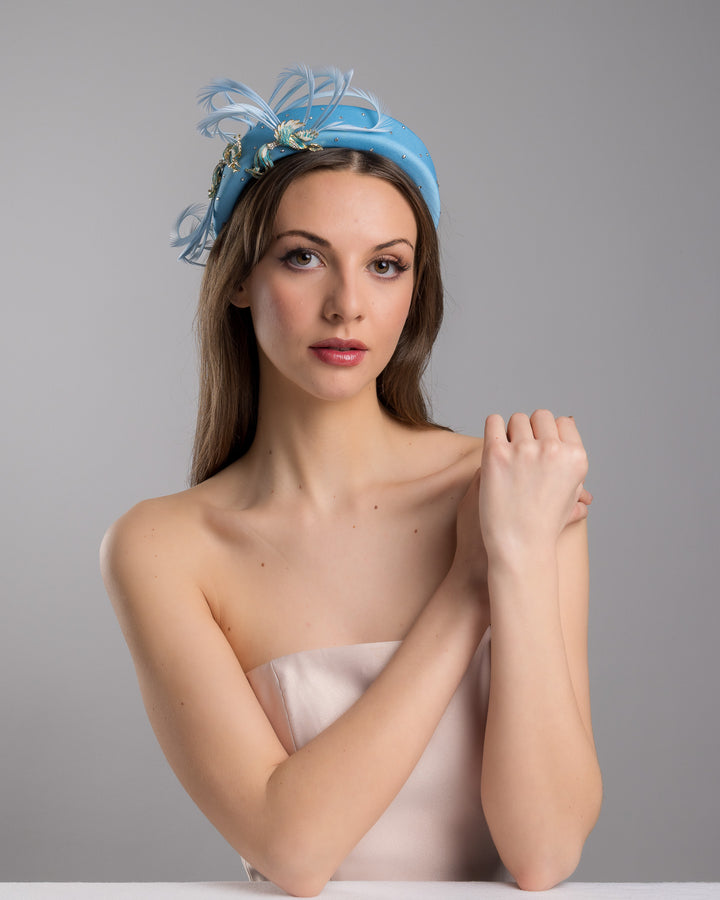 The female model is posing, wearing
Anqa, the aqua blue padded headband embellished with crystal accents, featuring gold and blue bird appliqués with flowing feather details for a bold, statement finish. Designed and made by Merve Bayindir Millinery.
