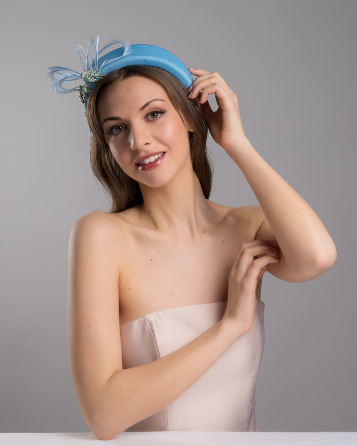 The female model is posing, wearing
Anqa, the aqua blue padded headband embellished with crystal accents, featuring gold and blue bird appliqués with flowing feather details for a bold, statement finish. Designed and made by Merve Bayindir Millinery.
