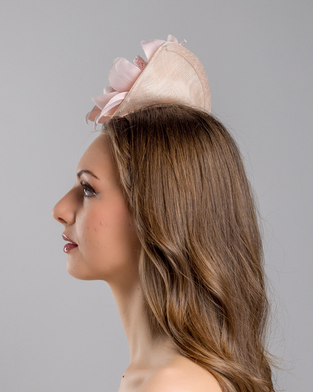 The model is showcasing the left side close up of the blush pink sinamay fascinator adorned with oversized handcrafted satin flowers, beaded centers, sculpted leaves, and curved stem details, mounted on a slim headband. Designed and made by Merve Bayindir Millinery.

