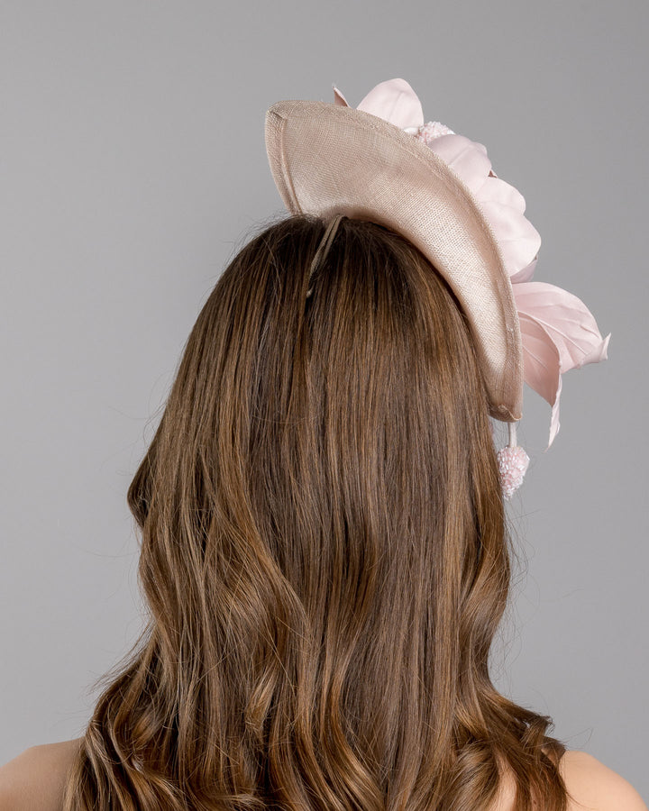 The model is showcasing the back side close up of the blush pink sinamay fascinator adorned with oversized handcrafted satin flowers, beaded centers, sculpted leaves, and curved stem details, mounted on a slim headband. Designed and made by Merve Bayindir Millinery.
