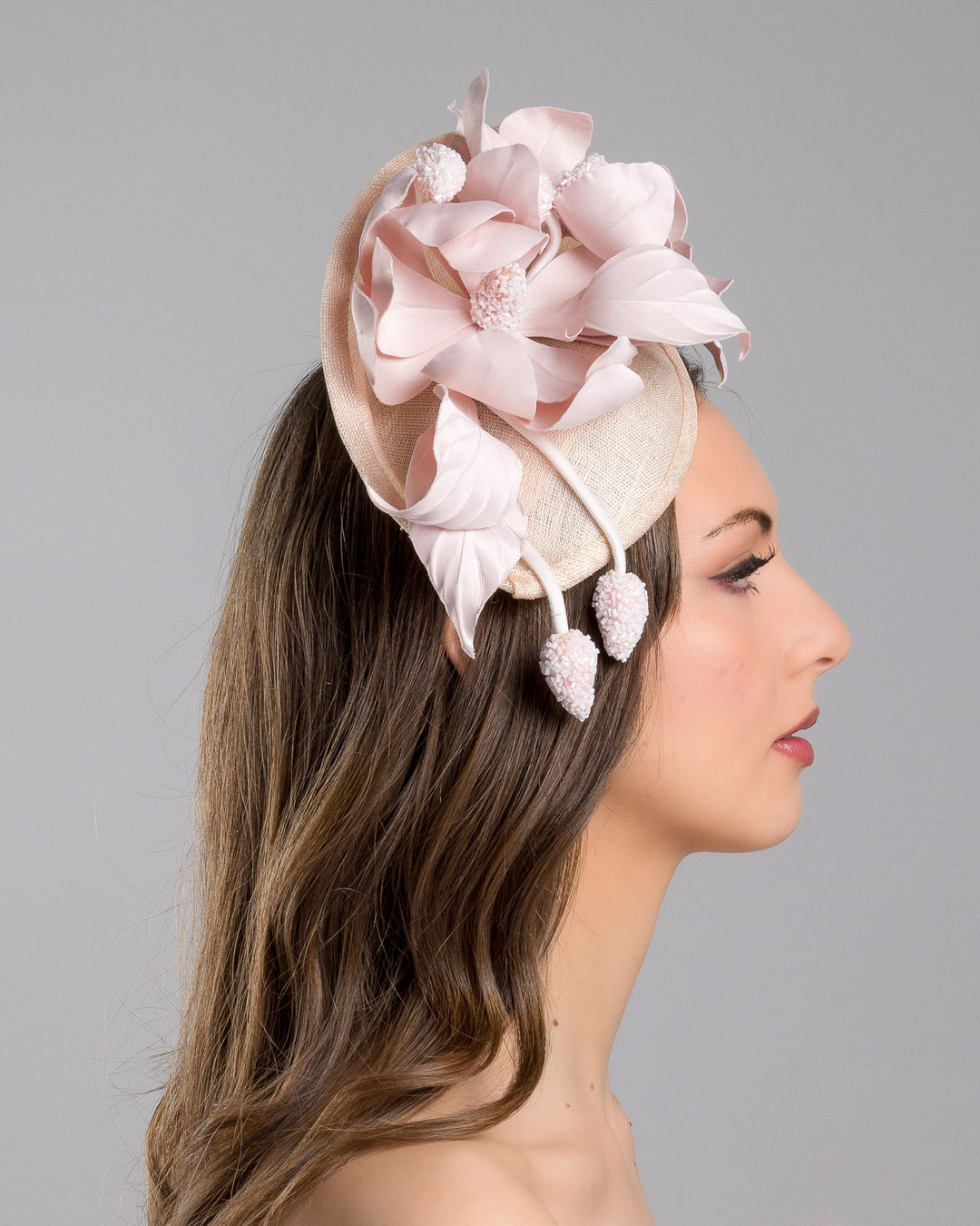 The model is showcasing the right side close up of the blush pink sinamay fascinator adorned with oversized handcrafted satin flowers, beaded centers, sculpted leaves, and curved stem details, mounted on a slim headband. Designed and made by Merve Bayindir Millinery.

