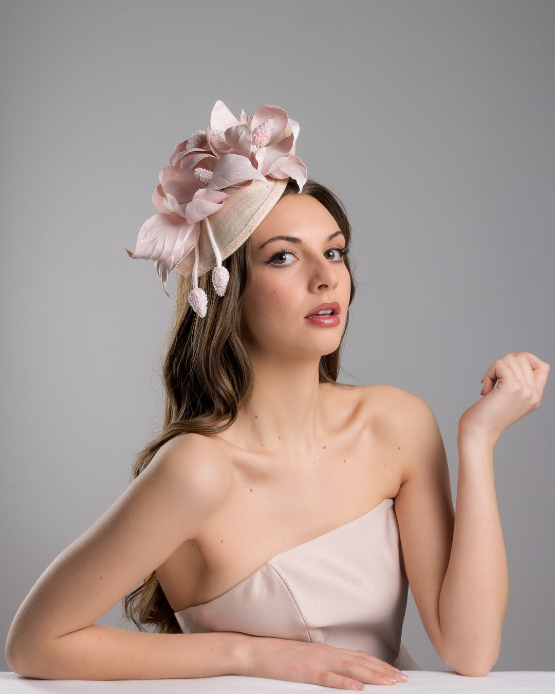 The female model is posing, wearing
Aphrodite, the blush pink sinamay fascinator adorned with oversized handcrafted satin flowers, beaded centers, sculpted leaves, and curved stem details, mounted on a slim headband. Designed and made by Merve Bayindir Millinery.
