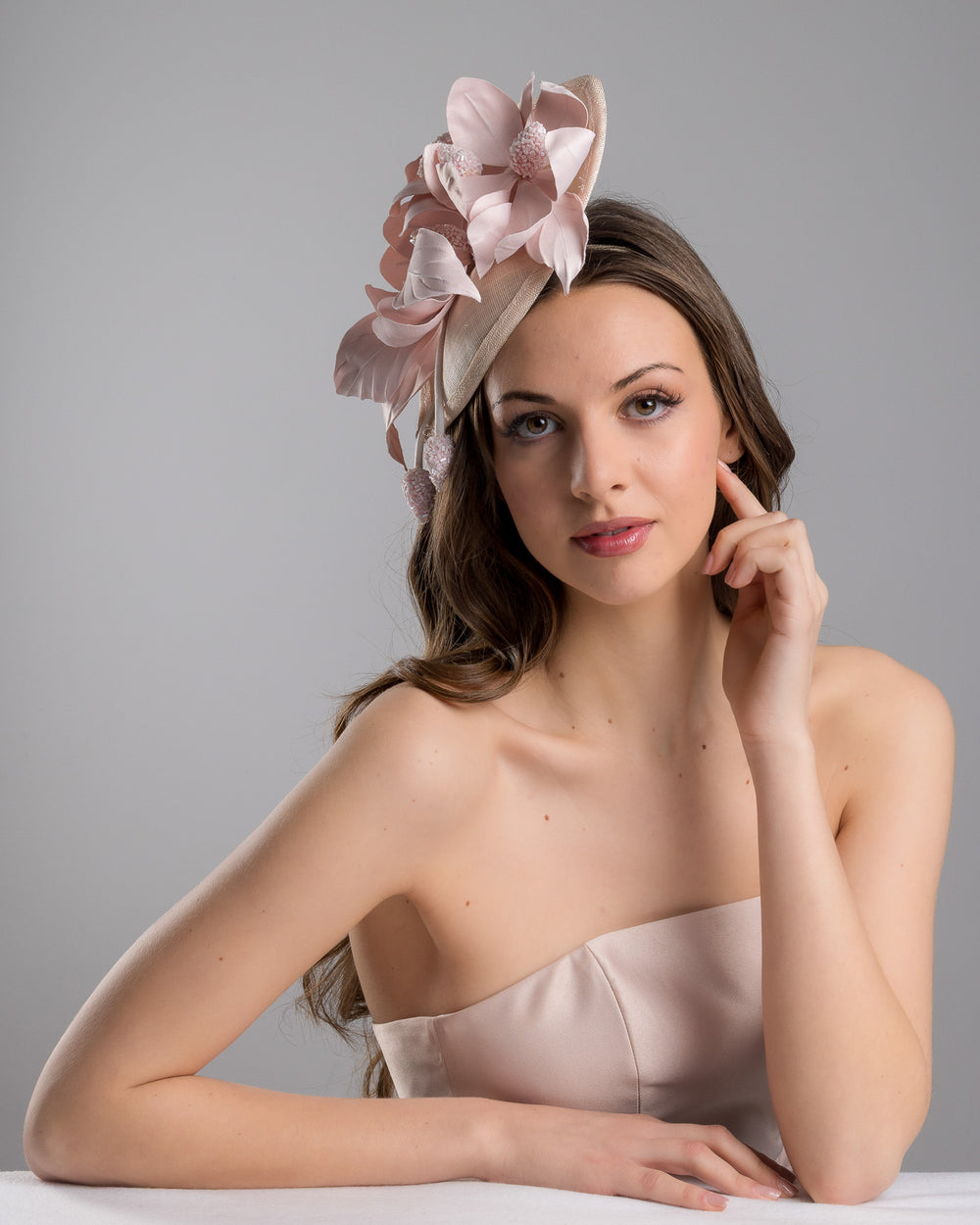 The female model is posing, wearing
Aphrodite, the blush pink sinamay fascinator adorned with oversized handcrafted satin flowers, beaded centers, sculpted leaves, and curved stem details, mounted on a slim headband. Designed and made by Merve Bayindir Millinery.
