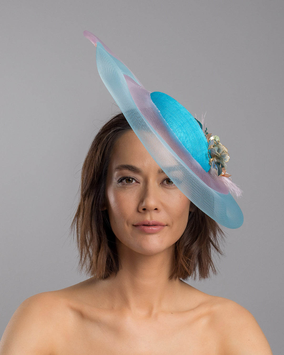 The model is showcasing the front side close up of the turquoise sinamay center base fascinator framed by concentric white and pale aqua sinamay rings, adorned with soft pastel feathers and delicate floral embellishments for a light, ethereal finish. Designed and made by Merve Bayindir Millinery.
