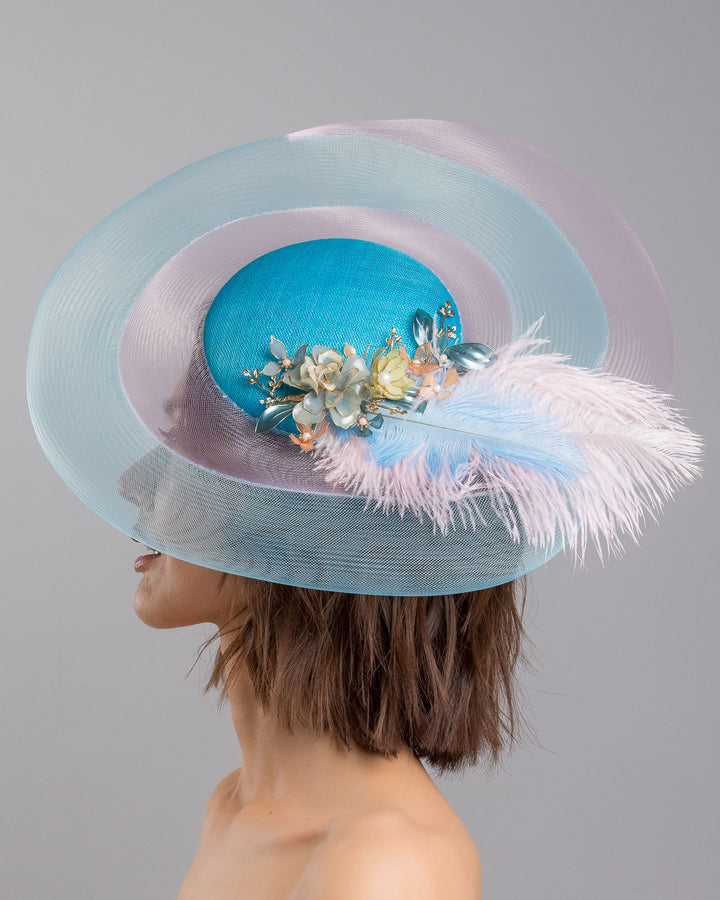 The model is showcasing the left side close up of the turquoise sinamay center base fascinator framed by concentric white and pale aqua sinamay rings, adorned with soft pastel feathers and delicate floral embellishments for a light, ethereal finish. Designed and made by Merve Bayindir Millinery.
