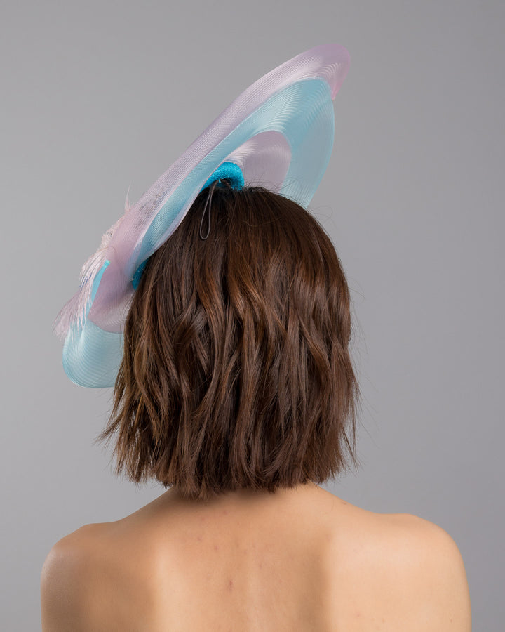 The model is showcasing the back side close up of the turquoise sinamay center base fascinator framed by concentric white and pale aqua sinamay rings, adorned with soft pastel feathers and delicate floral embellishments for a light, ethereal finish. Designed and made by Merve Bayindir Millinery.
