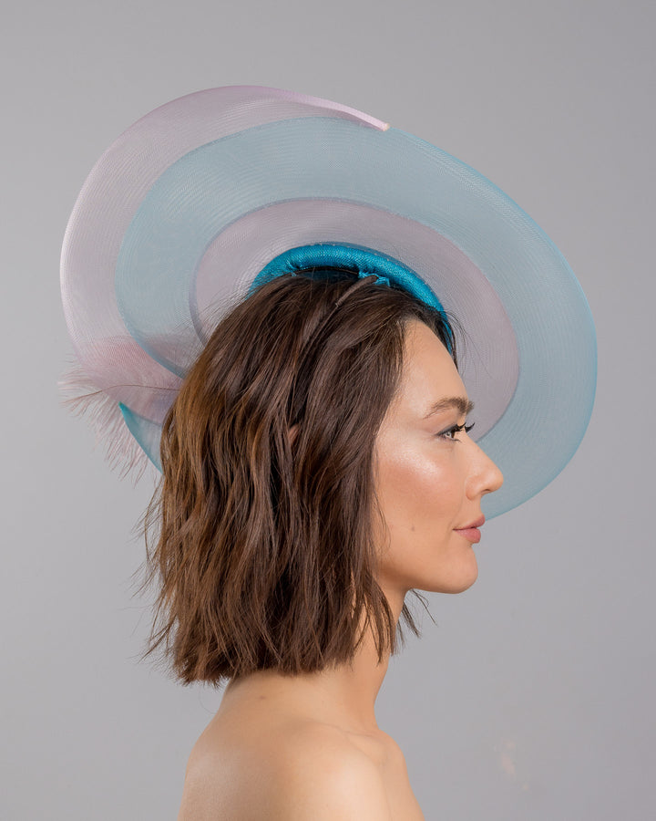 The model is showcasing the right side close up of the turquoise sinamay center base fascinator framed by concentric white and pale aqua sinamay rings, adorned with soft pastel feathers and delicate floral embellishments for a light, ethereal finish. Designed and made by Merve Bayindir Millinery.


