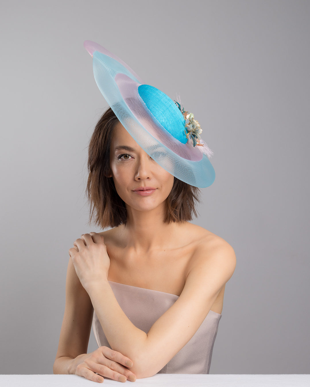 The female model is posing, wearing
Aquael, the turquoise sinamay center base fascinator framed by concentric white and pale aqua sinamay rings, adorned with soft pastel feathers and delicate floral embellishments for a light, ethereal finish. Designed and made by Merve Bayindir Millinery.
