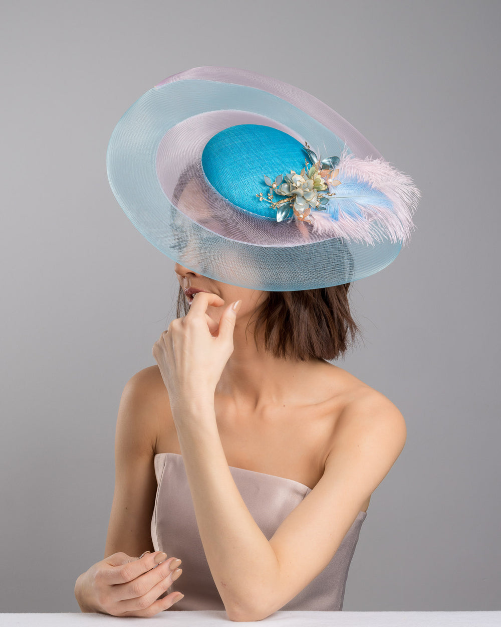 The female model is posing, wearing
Aquael, the turquoise sinamay center base fascinator framed by concentric white and pale aqua sinamay rings, adorned with soft pastel feathers and delicate floral embellishments for a light, ethereal finish. Designed and made by Merve Bayindir Millinery.

