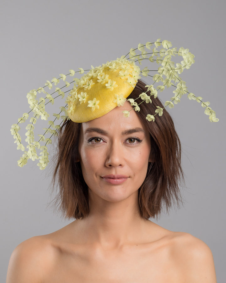 The model is showcasing the front side close up of the bright yellow sinamay base fascinator shaped in a smooth rounded form, adorned with clusters of small soft-yellow flowers and framed by airy trailing floral stems that curve gracefully around the piece. Designed and made by Merve Bayindir Millinery.