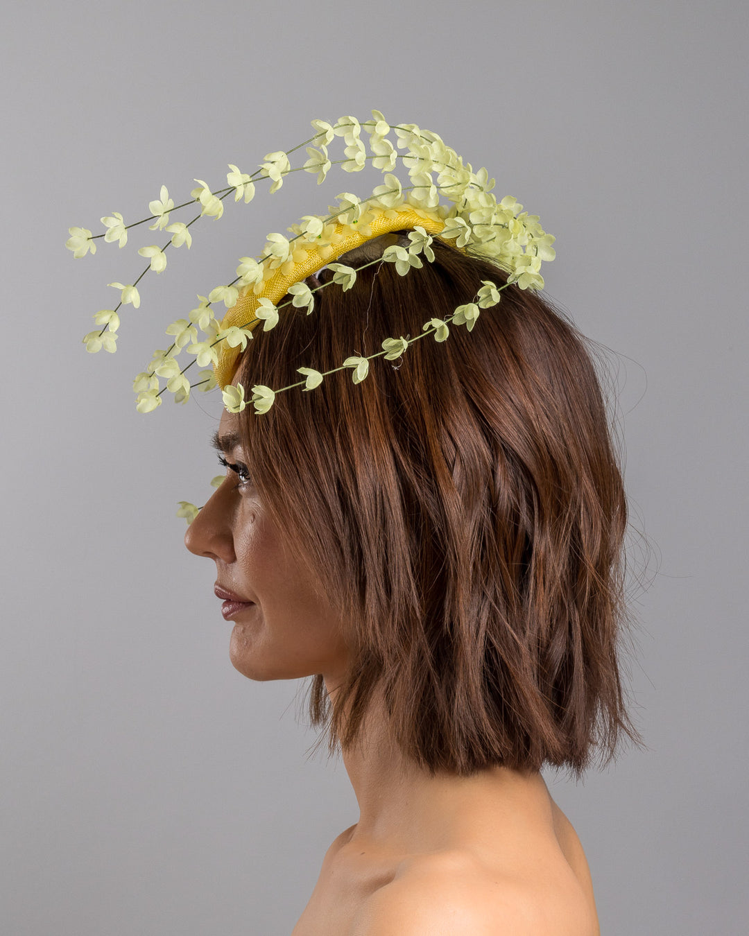 The model is showcasing the left side close up of the bright yellow sinamay base fascinator shaped in a smooth rounded form, adorned with clusters of small soft-yellow flowers and framed by airy trailing floral stems that curve gracefully around the piece. Designed and made by Merve Bayindir Millinery.