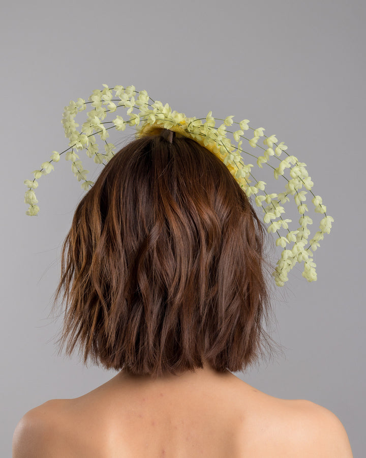 The model is showcasing the back side close up of the bright yellow sinamay base fascinator shaped in a smooth rounded form, adorned with clusters of small soft-yellow flowers and framed by airy trailing floral stems that curve gracefully around the piece. Designed and made by Merve Bayindir Millinery.