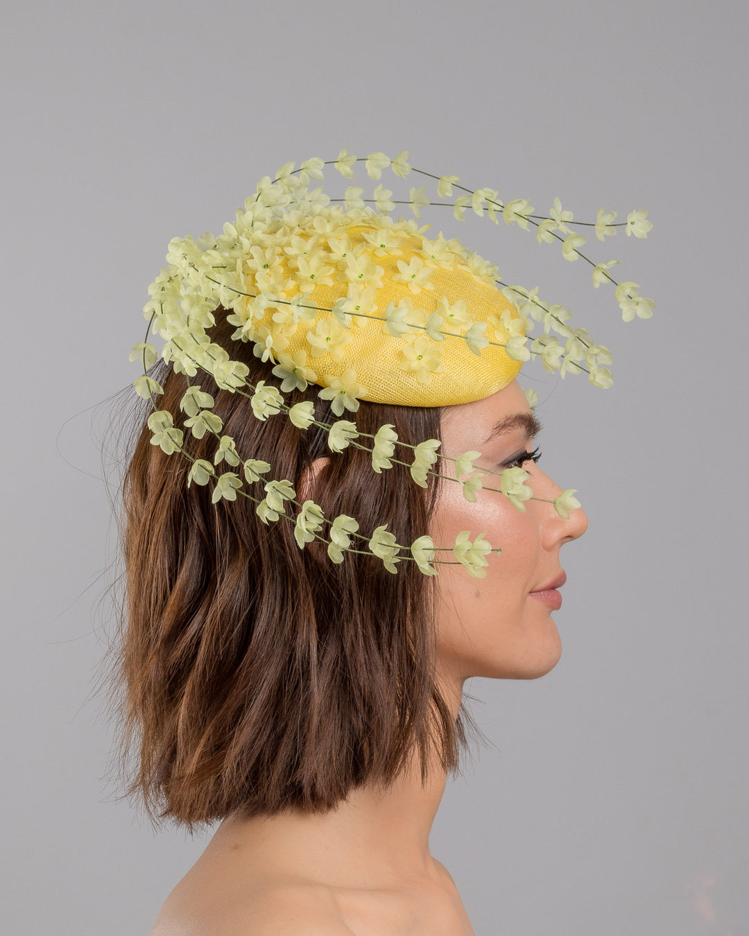 The model is showcasing the right side close up of the bright yellow sinamay base fascinator shaped in a smooth rounded form, adorned with clusters of small soft-yellow flowers and framed by airy trailing floral stems that curve gracefully around the piece. Designed and made by Merve Bayindir Millinery.
