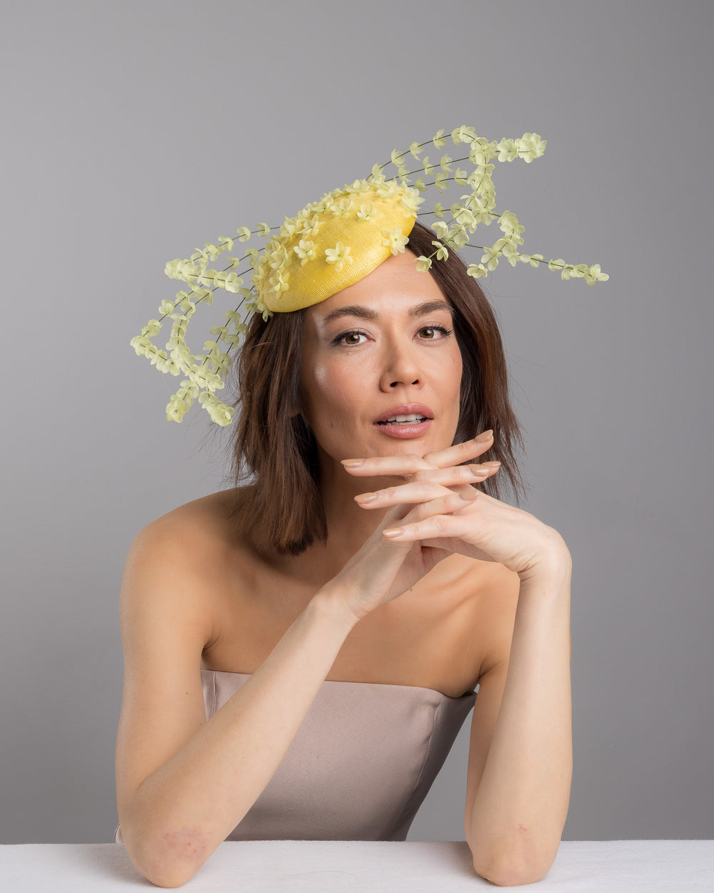 The female model is posing, wearing
Ardena, the bright yellow sinamay base fascinator shaped in a smooth rounded form, adorned with clusters of small soft-yellow flowers and framed by airy trailing floral stems that curve gracefully around the piece. Designed and made by Merve Bayindir Millinery.