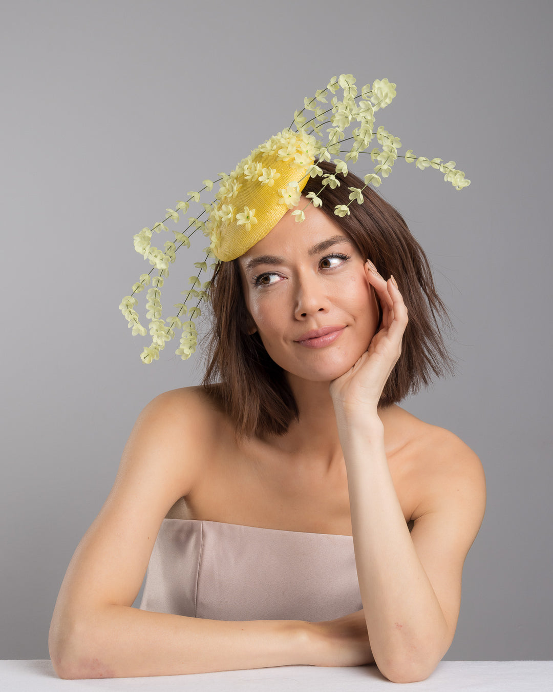 The female model is posing, wearing
Ardena, the bright yellow sinamay base fascinator shaped in a smooth rounded form, adorned with clusters of small soft-yellow flowers and framed by airy trailing floral stems that curve gracefully around the piece. Designed and made by Merve Bayindir Millinery.