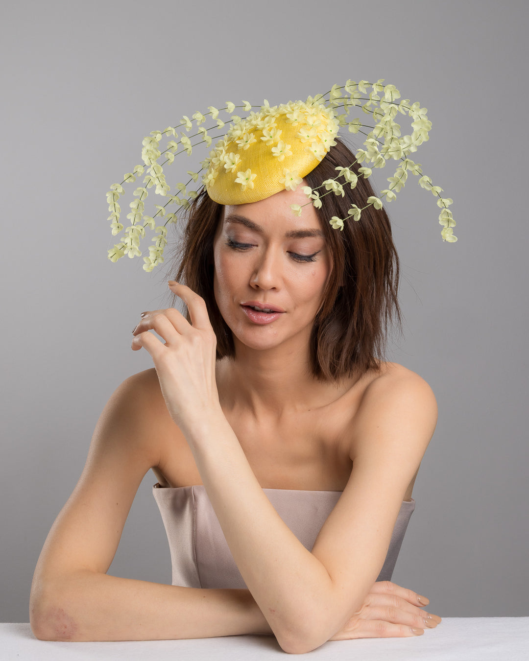 The female model is posing, wearing
Ardena, the bright yellow sinamay base fascinator shaped in a smooth rounded form, adorned with clusters of small soft-yellow flowers and framed by airy trailing floral stems that curve gracefully around the piece. Designed and made by Merve Bayindir Millinery.