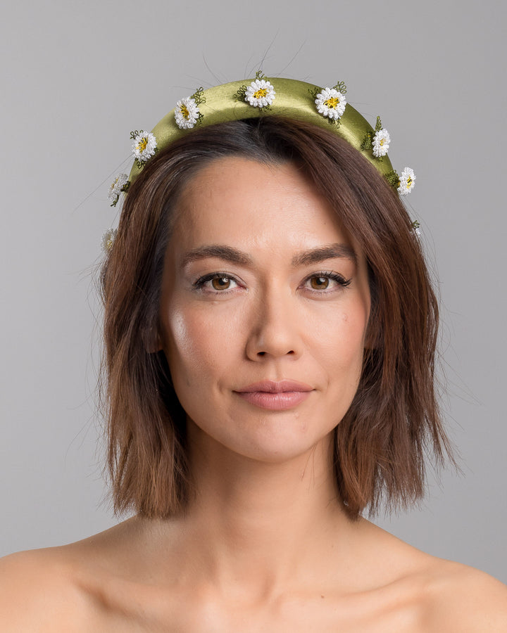 The model is showcasing the front side close up of the olive green satin padded headband embellished with small hand embroidered white daisy flowers featuring yellow centres and delicate green thread leaves spaced around the band. Designed and made by Merve Bayindir Millinery.
