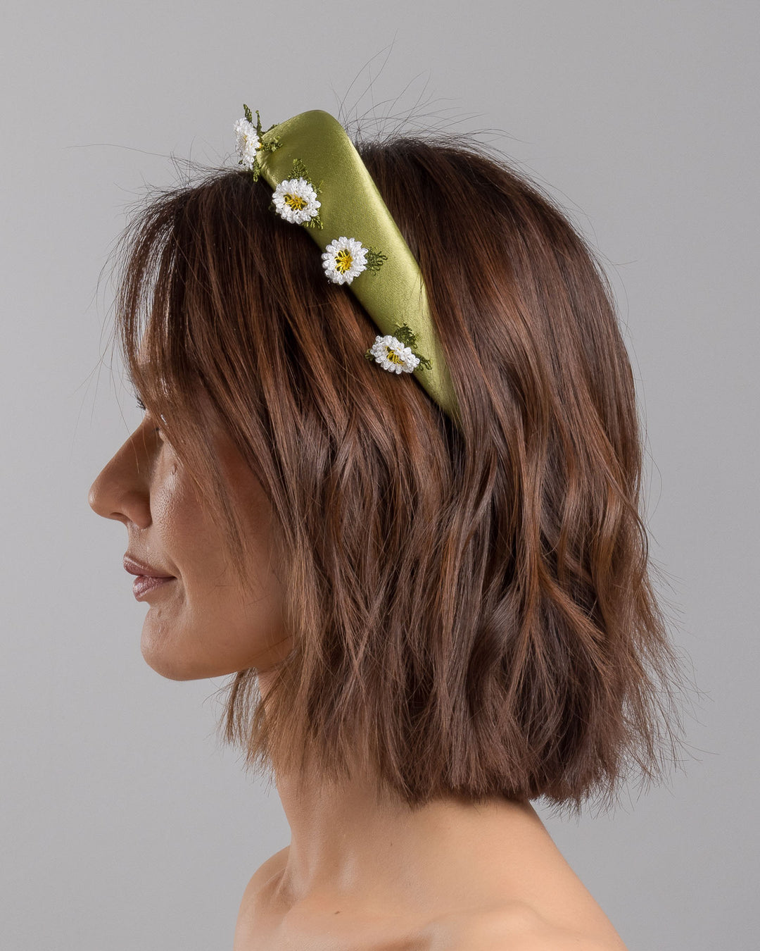 The model is showcasing the left side close up of the olive green satin padded headband embellished with small hand embroidered white daisy flowers featuring yellow centres and delicate green thread leaves spaced around the band. Designed and made by Merve Bayindir Millinery.
