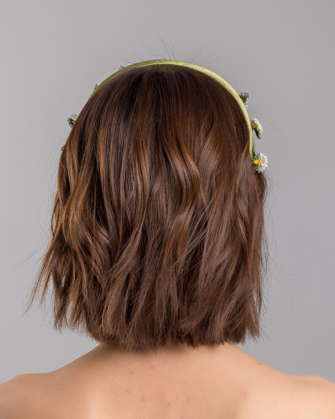 The model is showcasing the back side close up of the olive green satin padded headband embellished with small hand embroidered white daisy flowers featuring yellow centres and delicate green thread leaves spaced around the band. Designed and made by Merve Bayindir Millinery.
