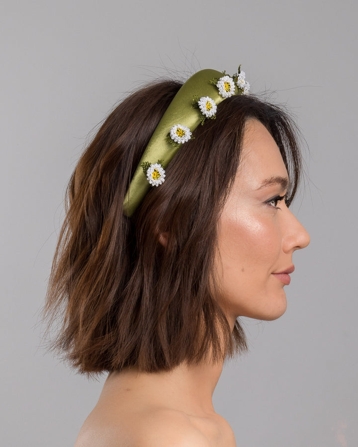 The model is showcasing the right side close up of the olive green satin padded headband embellished with small hand embroidered white daisy flowers featuring yellow centres and delicate green thread leaves spaced around the band. Designed and made by Merve Bayindir Millinery.

