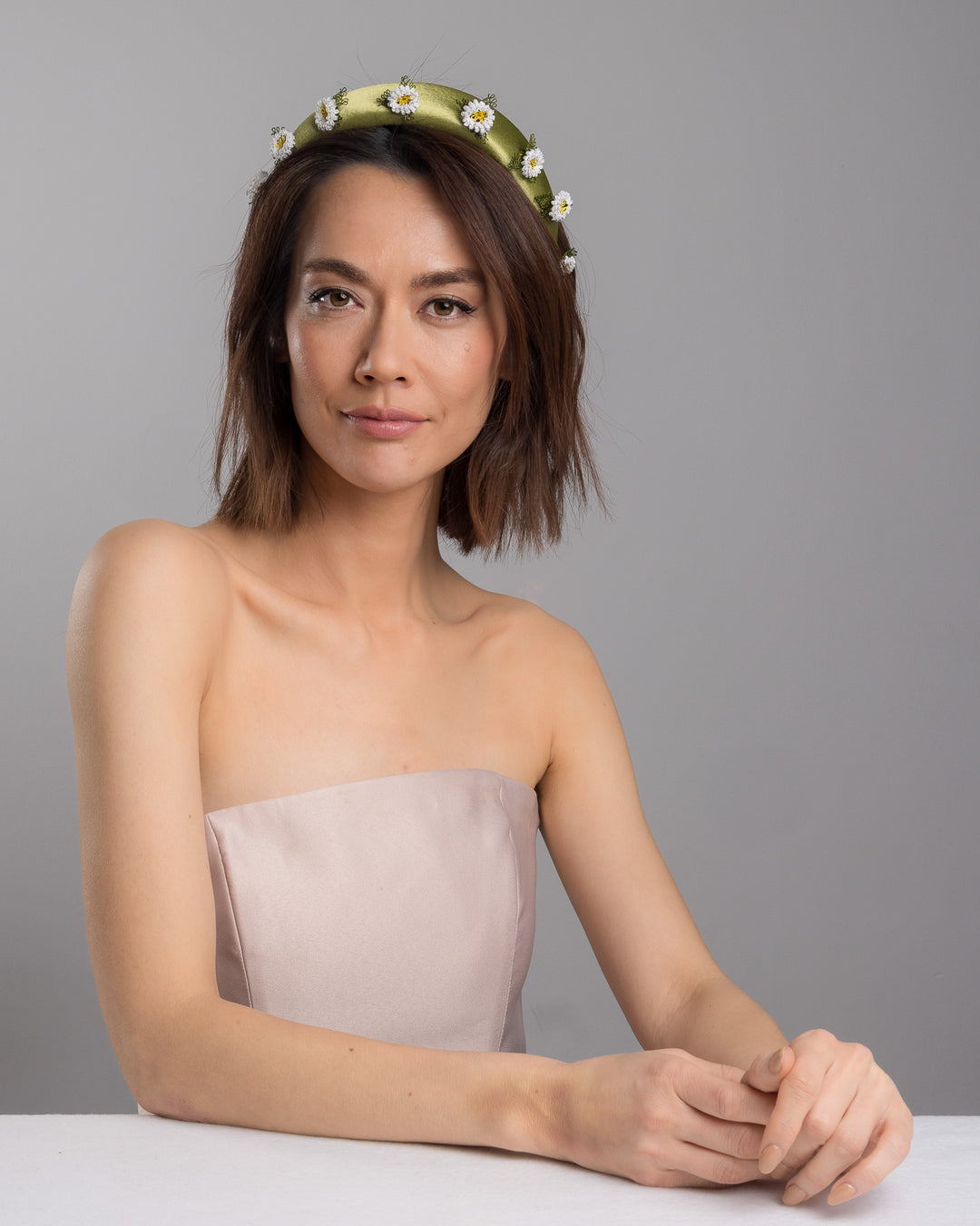 The female model is posing, wearing Arwenne, the olive green satin padded headband embellished with small hand embroidered white daisy flowers featuring yellow centres and delicate green thread leaves spaced around the band. Designed and made by Merve Bayindir Millinery.
