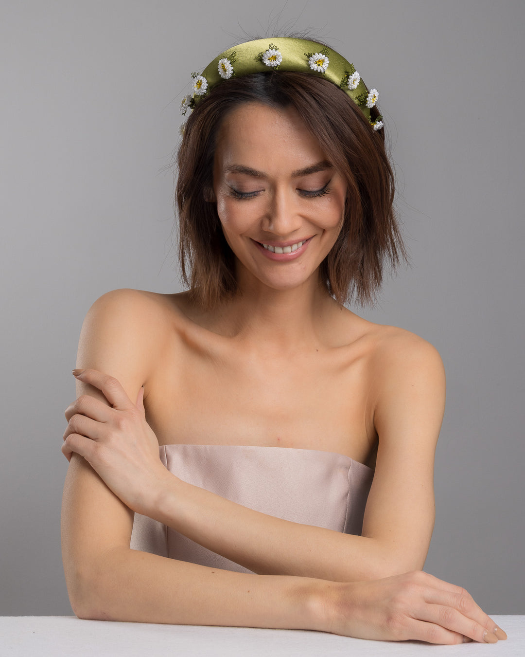 The female model is posing, wearing Arwenne, the olive green satin padded headband embellished with small hand embroidered white daisy flowers featuring yellow centres and delicate green thread leaves spaced around the band. Designed and made by Merve Bayindir Millinery.
