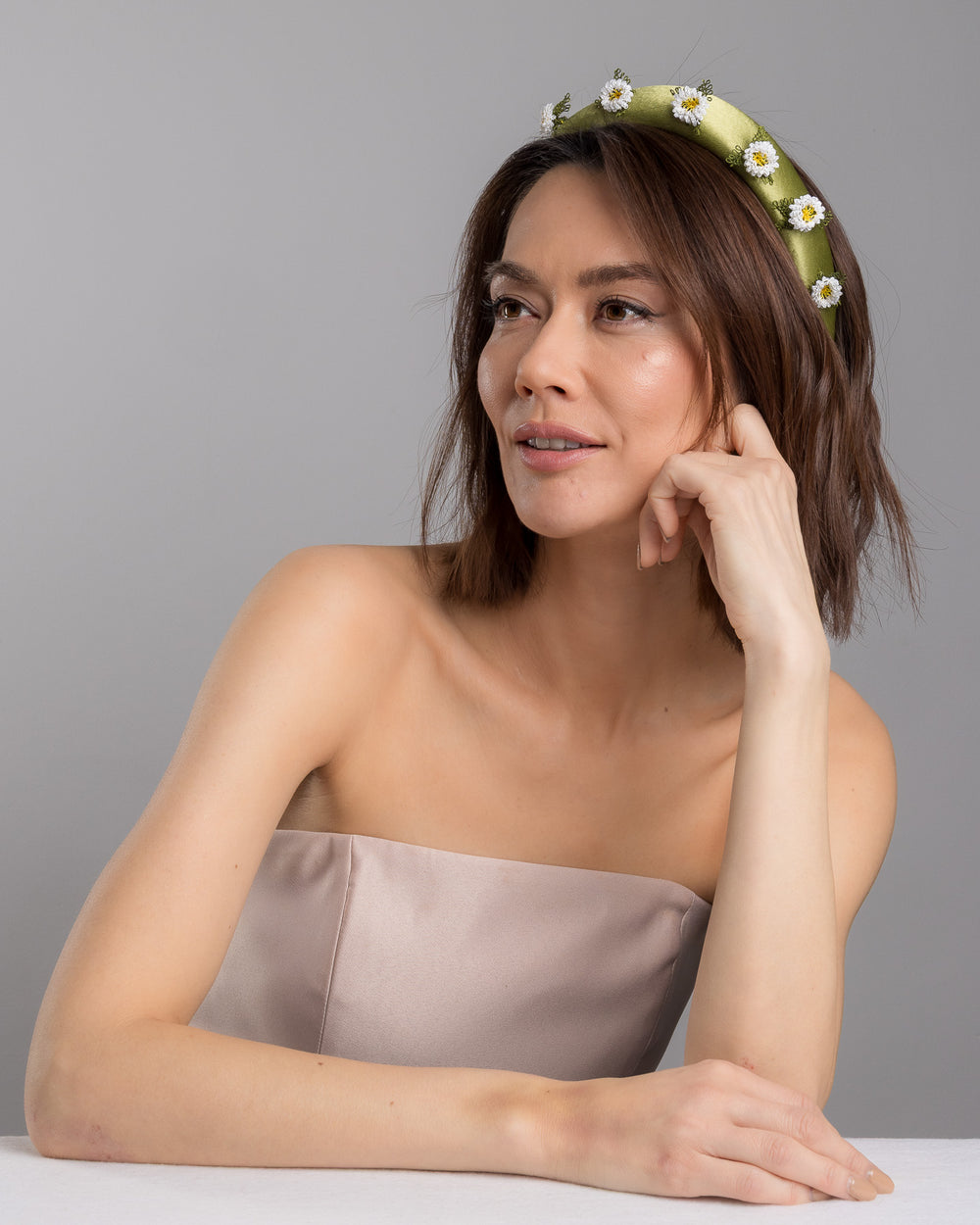 The female model is posing, wearing Arwenne, the olive green satin padded headband embellished with small hand embroidered white daisy flowers featuring yellow centres and delicate green thread leaves spaced around the band. Designed and made by Merve Bayindir Millinery.
