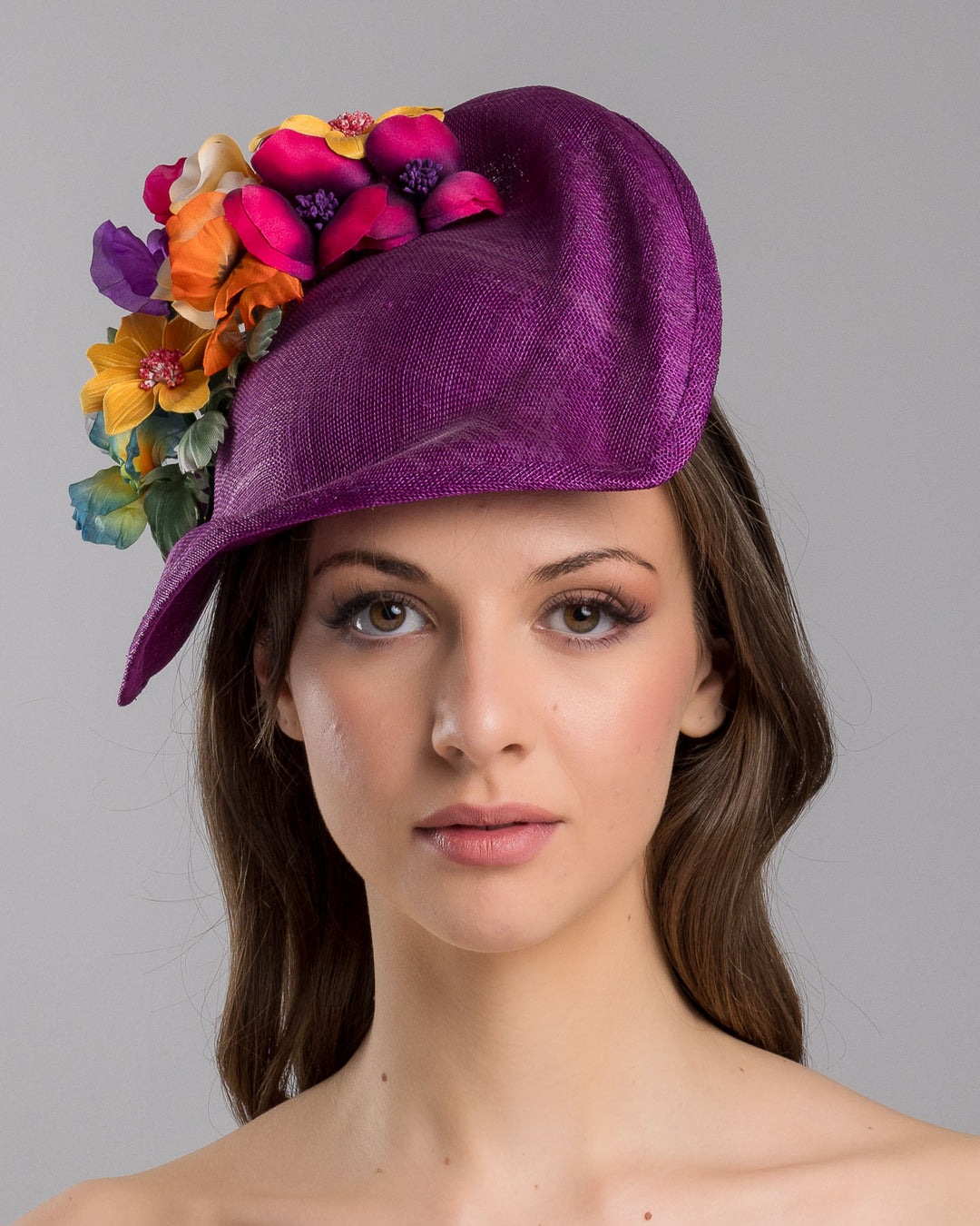 The model is showcasing the front side close up of the purple sinamay sculpted fascinator with a curved, wave-like shape, adorned with a vibrant cluster of multicolored flowers and mounted on a slim black headband. Designed and made by Merve Bayindir Millinery.
