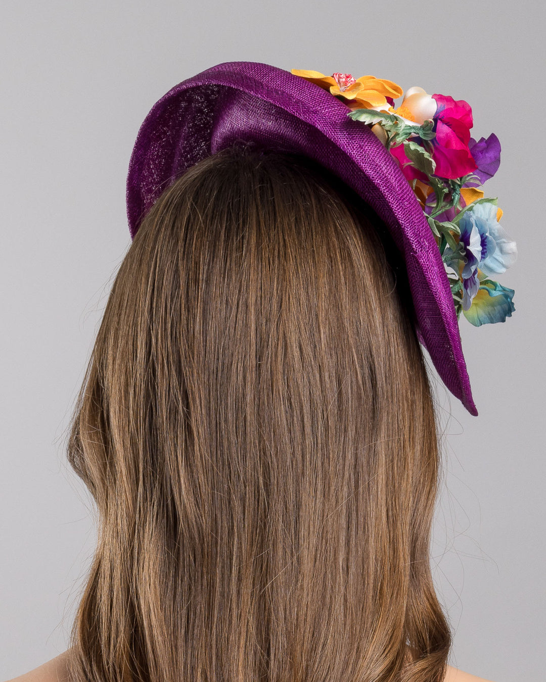 The model is showcasing the back side close up of the purple sinamay sculpted fascinator with a curved, wave-like shape, adorned with a vibrant cluster of multicolored flowers and mounted on a slim black headband. Designed and made by Merve Bayindir Millinery.
