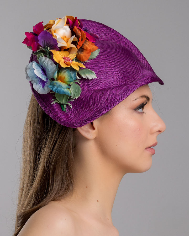 The model is showcasing the right side close up of the purple sinamay sculpted fascinator with a curved, wave-like shape, adorned with a vibrant cluster of multicolored flowers and mounted on a slim black headband. Designed and made by Merve Bayindir Millinery.
