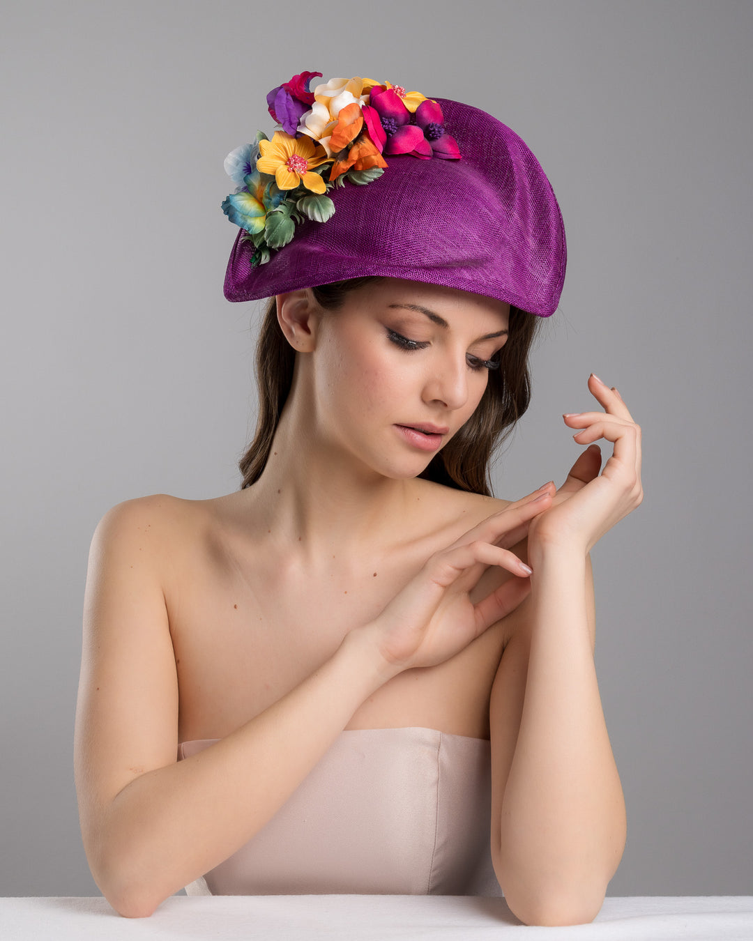 The female model is posing, wearing
Avery, the purple sinamay sculpted fascinator with a curved, wave-like shape, adorned with a vibrant cluster of multicolored flowers and mounted on a slim black headband. Designed and made by Merve Bayindir Millinery.
