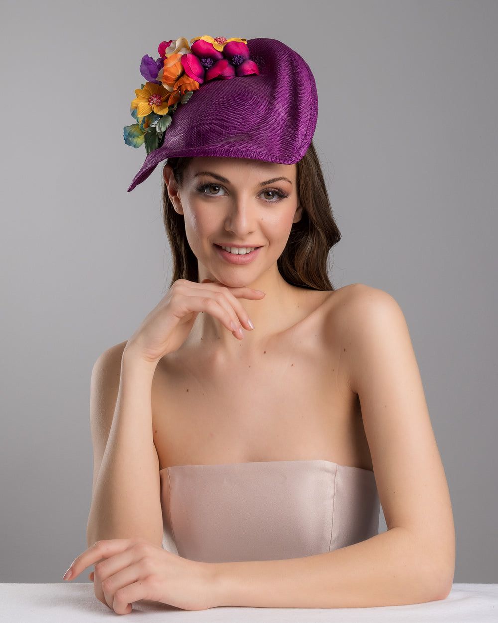 The female model is posing, wearing
Avery, the purple sinamay sculpted fascinator with a curved, wave-like shape, adorned with a vibrant cluster of multicolored flowers and mounted on a slim black headband. Designed and made by Merve Bayindir Millinery.

