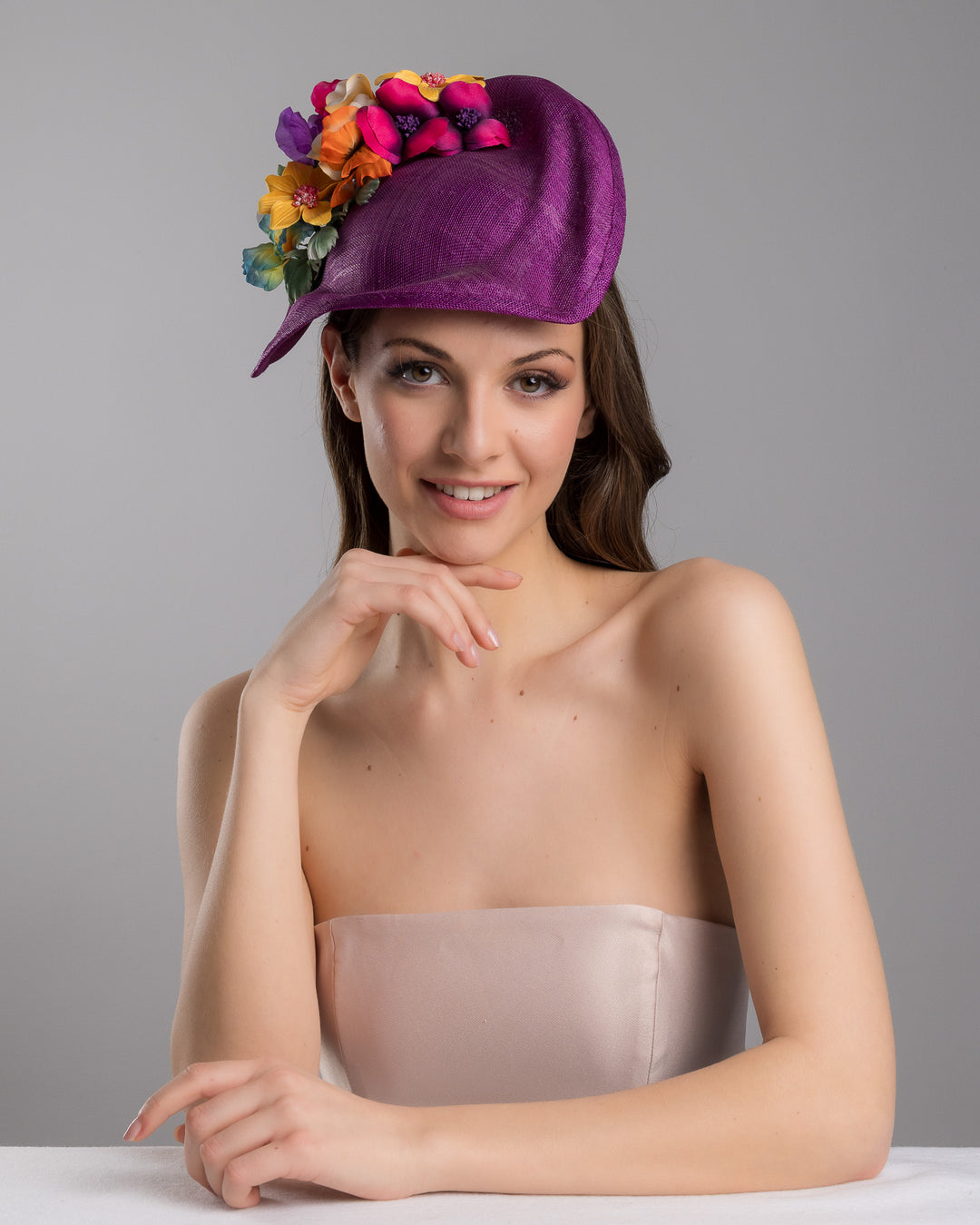 The female model is posing, wearing
Avery, the purple sinamay sculpted fascinator with a curved, wave-like shape, adorned with a vibrant cluster of multicolored flowers and mounted on a slim black headband. Designed and made by Merve Bayindir Millinery.
