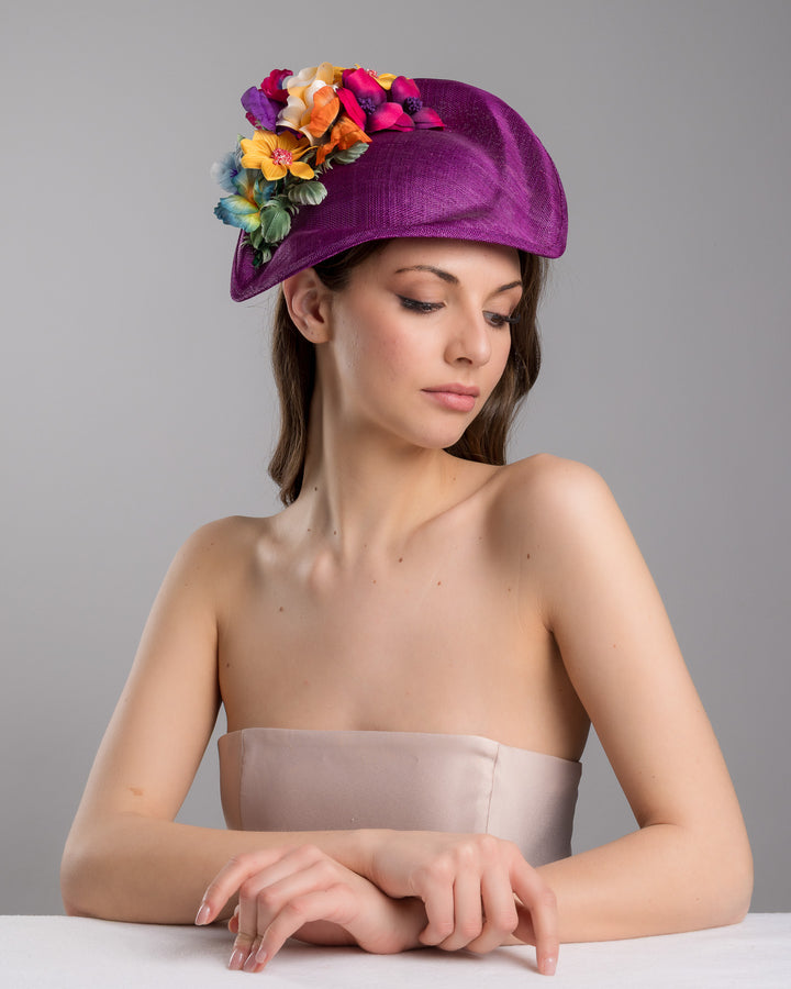 The female model is posing, wearing
Avery, the purple sinamay sculpted fascinator with a curved, wave-like shape, adorned with a vibrant cluster of multicolored flowers and mounted on a slim black headband. Designed and made by Merve Bayindir Millinery.
