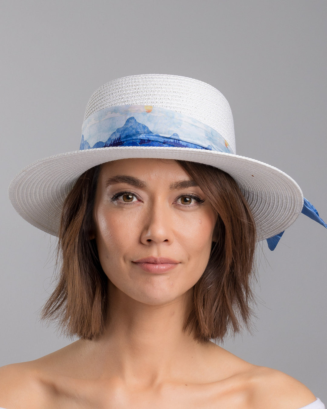 The model is showcasing the front side close up of the white woven straw boater hat with a wide brim, wrapped in a soft blue watercolour scarf and finished with delicate blue beaded floral embellishments at the side. Designed by Merve Bayindir millinery.