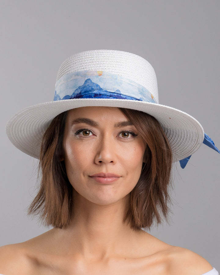 The model is showcasing the front side close up of the white woven straw boater hat with a wide brim, wrapped in a soft blue watercolour scarf and finished with delicate blue beaded floral embellishments at the side. Designed by Merve Bayindir millinery.