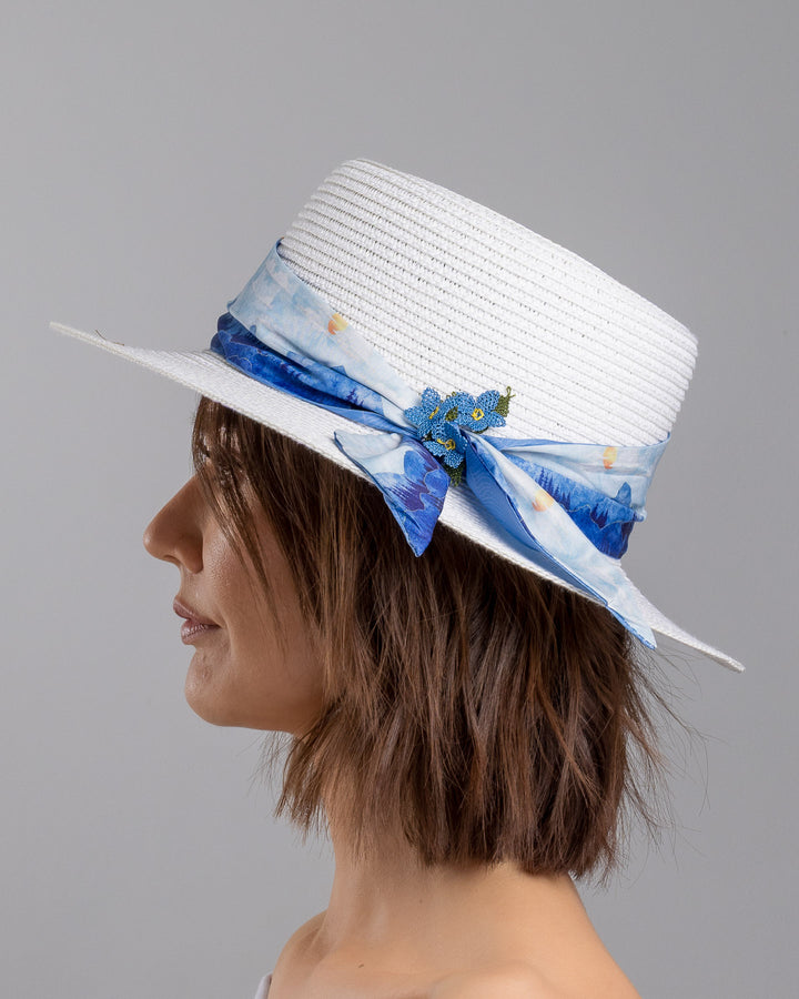 The model is showcasing the left side close up of the white woven straw boater hat with a wide brim, wrapped in a soft blue watercolour scarf and finished with delicate blue beaded floral embellishments at the side. Designed by Merve Bayindir millinery.